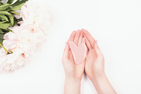 Pink flowers on white background near woman's hand holding pink, heart-shaped gua sha stone. 