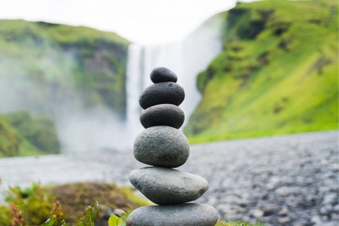 Smooth, round stones stacked 6-high in front of a waterfall surrounded by green hills. 
